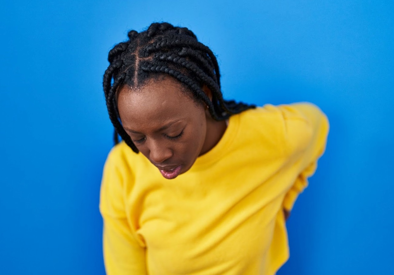 A black woman standing over a blue background suffering from lower back ache.