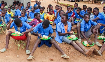 African school children sitting on an open ground and having a meal