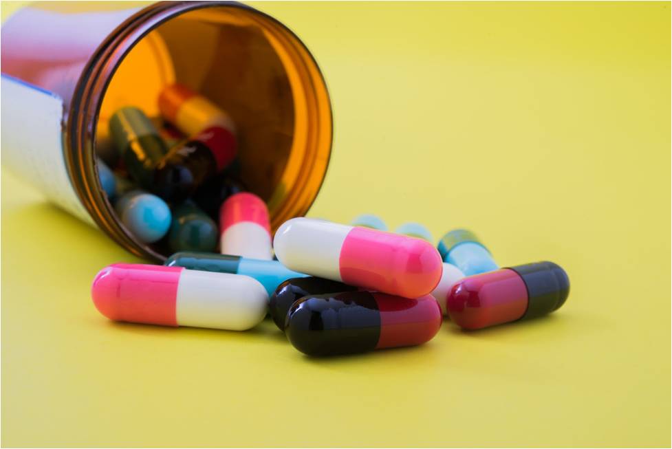 A tipped amber pill bottle with colorful capsules spilling onto a yellow background.