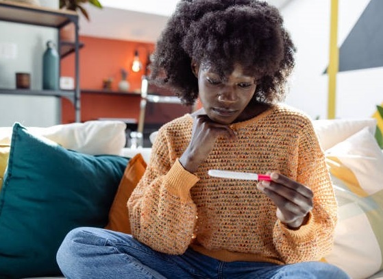 A black woman sitting on a couch observing a pregnancy test kit held in her left hand