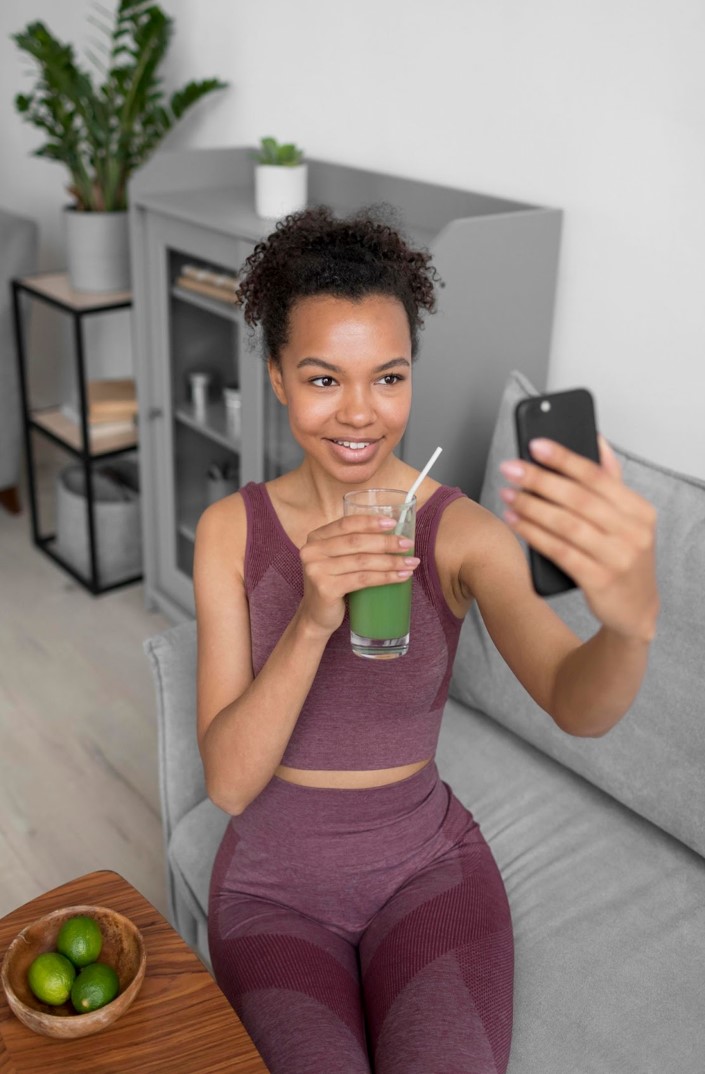 A young black lady sitting on a couch and taking a selfie with a glass of smoothie in her right hand