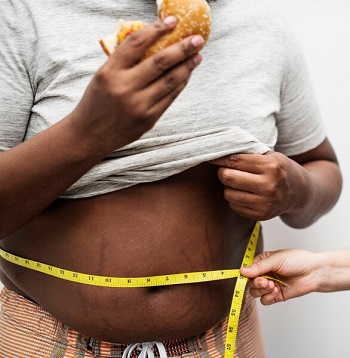 An obese man having his waistline measured while holding a hamburger in one hand