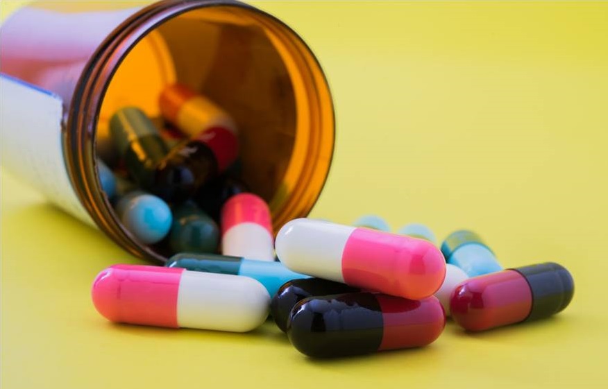 A tipped amber pill bottle with colorful capsules spilling onto a yellow background.