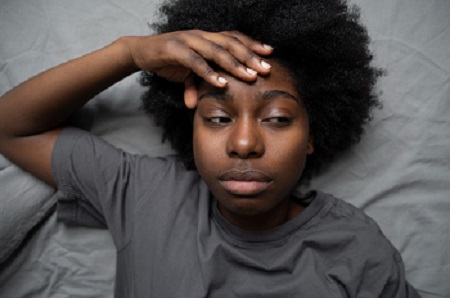 A young black lady lying in bed with hands on the head and eyes open.